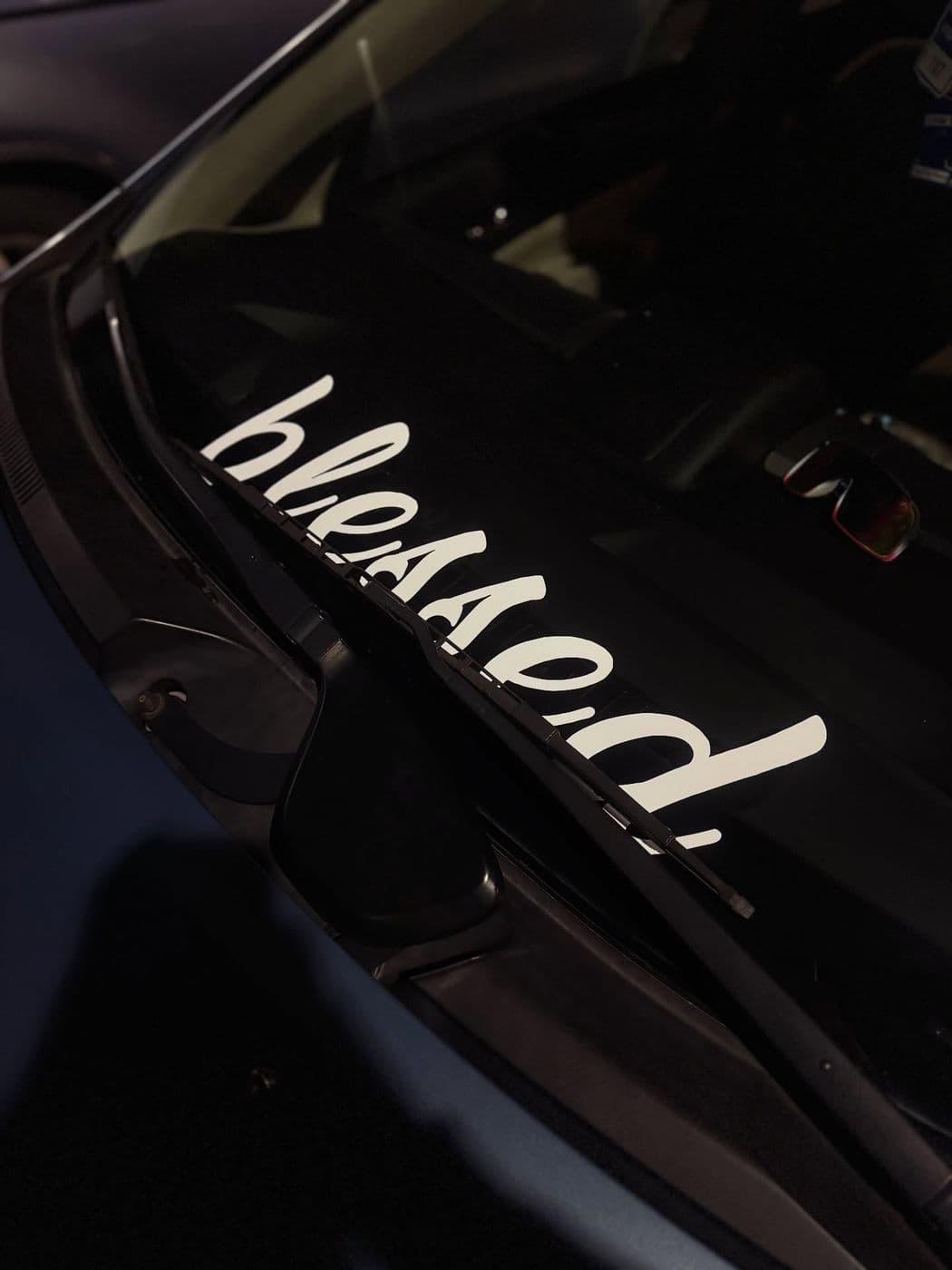 White script “blessed.” vinyl banner across a black car’s windshield